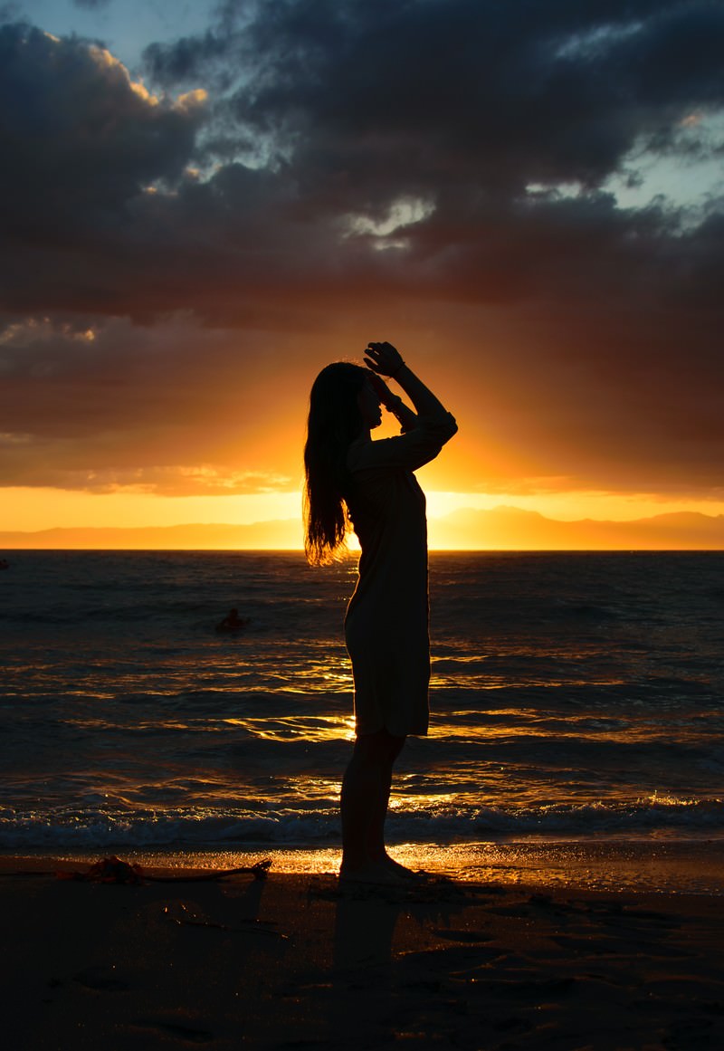 夕日をバックに砂浜で髪をかきあげようとする女性のシルエットの写真素材 ぱくたそ